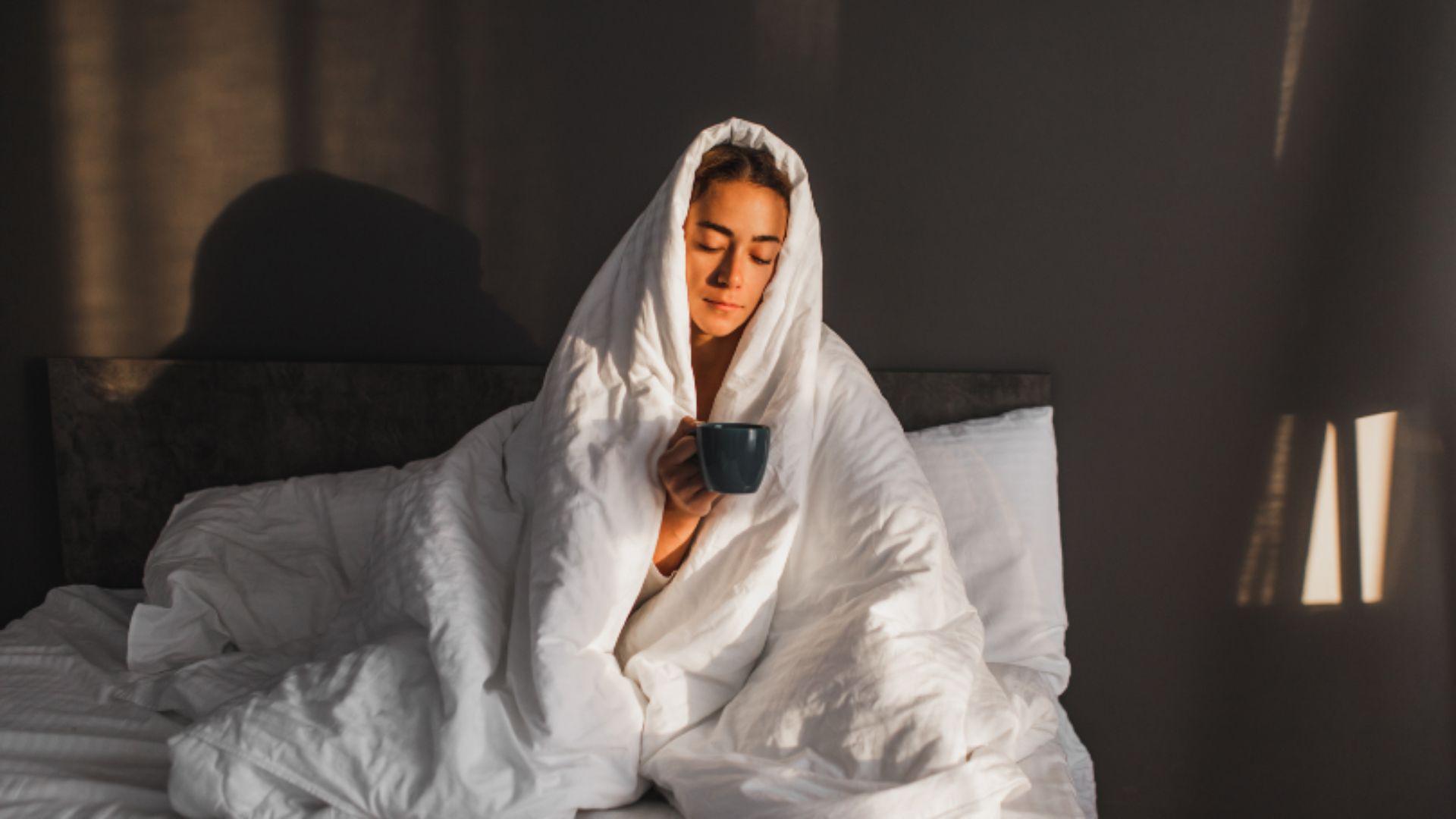 Young woman in bed with the duvet over her head, holding onto a mug.
