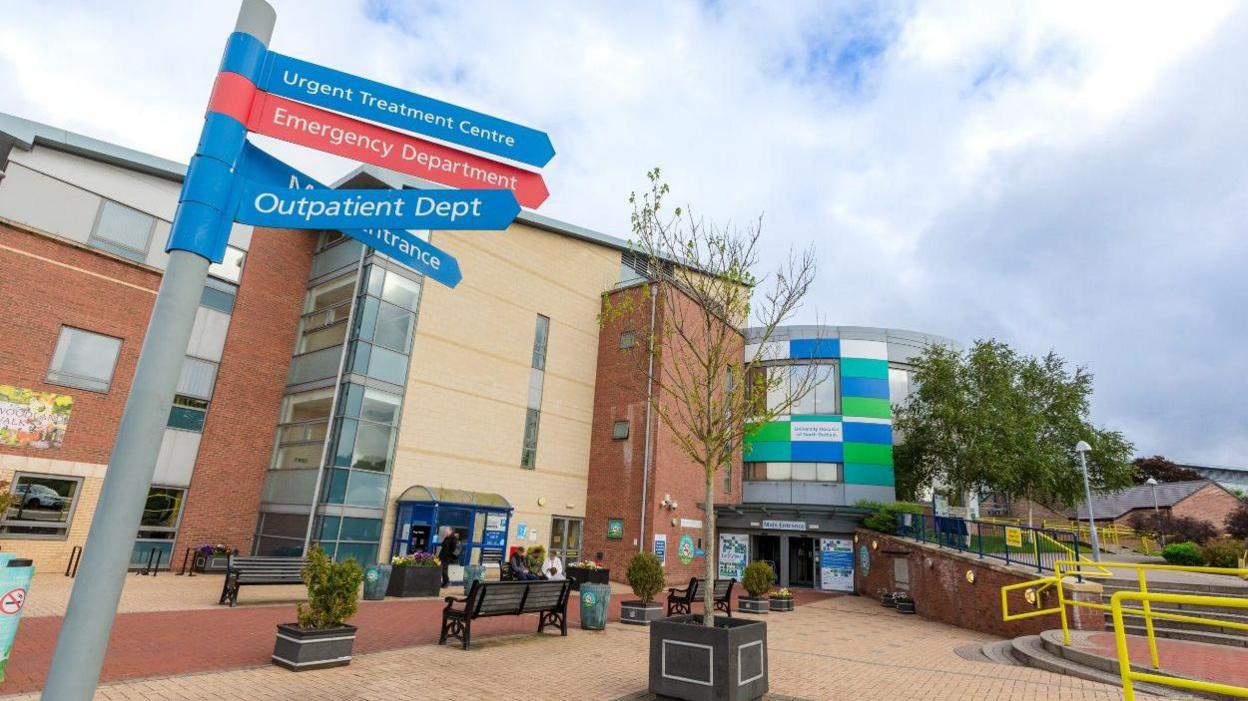 The outside of the University of North Durham Hospital. It a brown and beige-coloured brick building. The entrance has green, white and blue rectangle panelling. There is a sign pointing patients to various departments. A small tree is in a plant pot in a seating area.