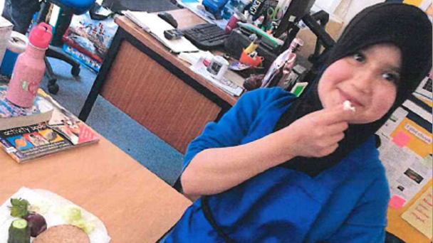 Sara Sharif wearing a black hijab and blue outfit. She appears to be in a classroom of office. She is holding a piece of food near to her mouth.