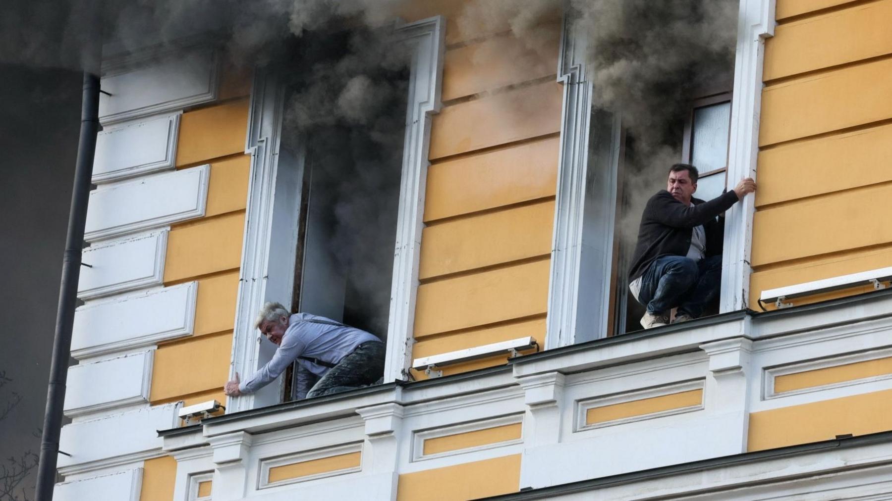Plume of black smoke coming out of window as two men lean out of windows