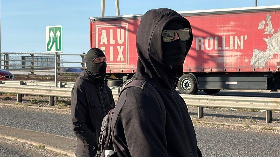 Two men wearing balaclavas stand on a road. There is a lorry in the background across the central reservation