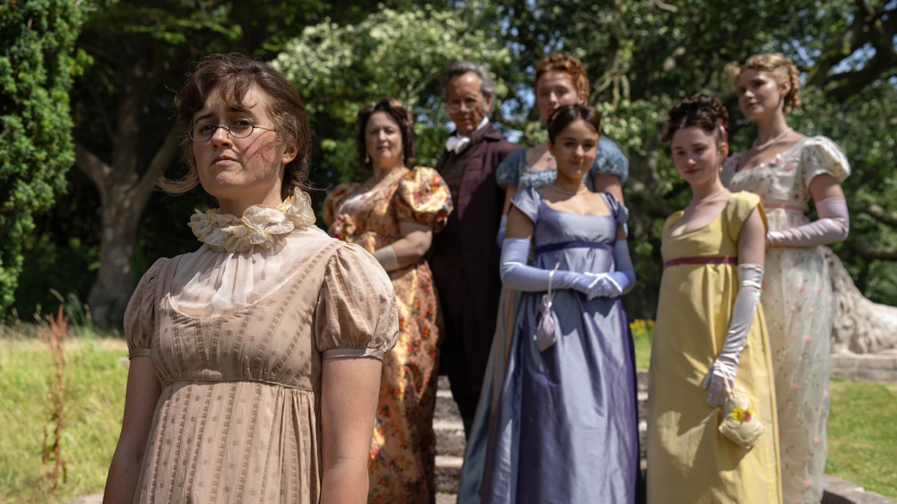 The family, except Mary, stand on steps outside in a garden. Mary stands closer to the camera, in a beige dress with a mesh high neck.