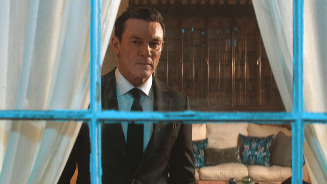 Evans is staring past the camera through a window with a chipped white frame. He wears a black suit and tie, with a white shirt.