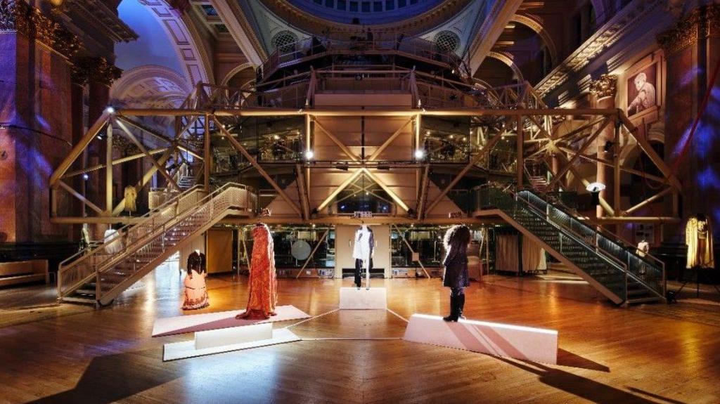 Costumes on display in the great hall of the Royal Exchange Theatre. Spotlights are shining on four different costumes from shows.