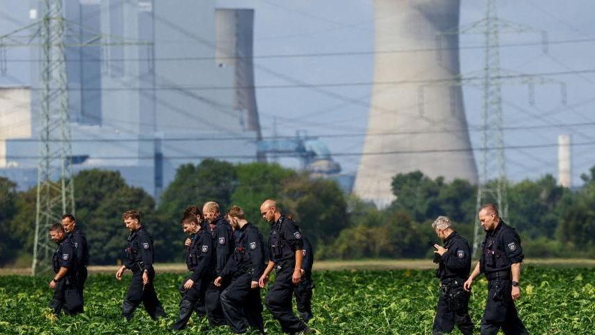About a dozen German police officers in black overalls are seen combing a field during a search. An active power station is seen in the background.