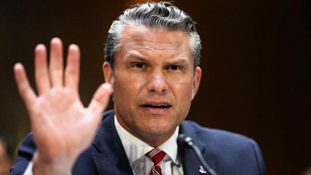 Pete Hegseth gestures by lifting his hand as he frowns