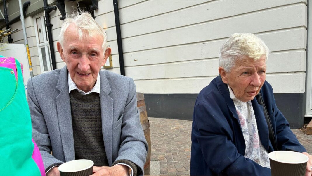 An elderly man in a grey suit and dark-coloured jumper. He is sitting next to an elderly woman wearing a dark blue parka and patterned jumper. Each has a cup of coffee in front of them.