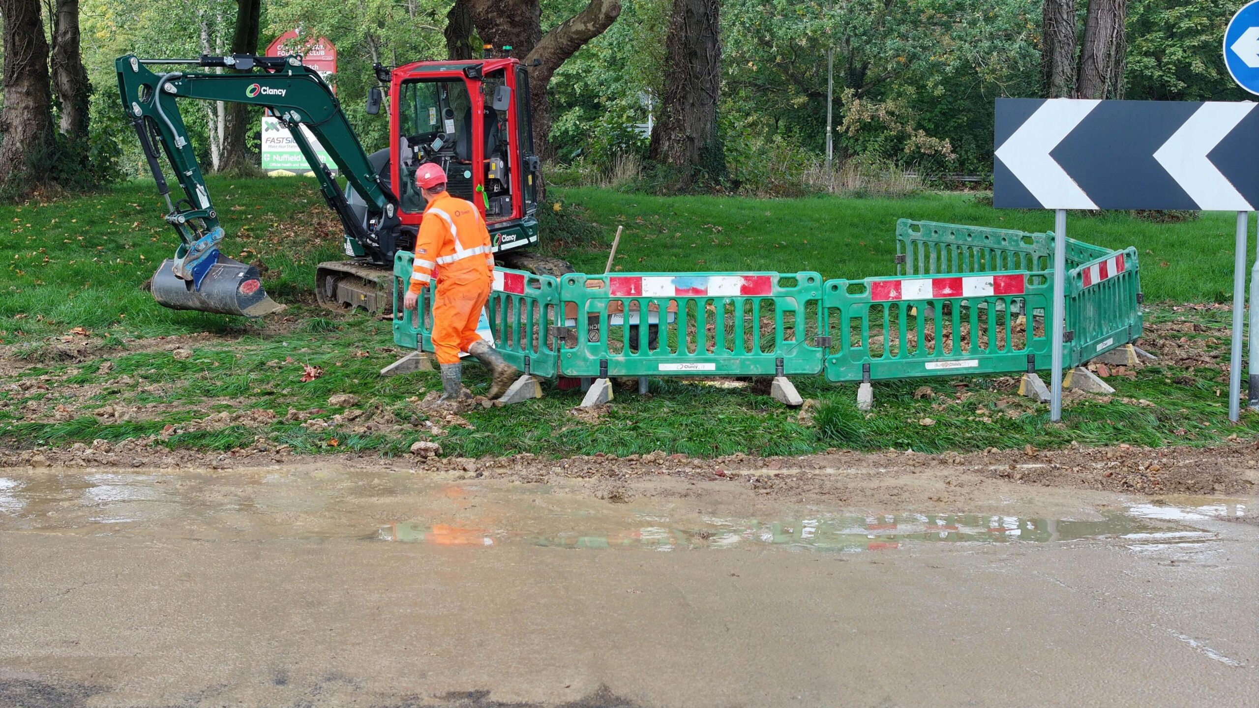 Workman in road wearing hi vis next to floods. The side of the road has a grassy bank and trees.