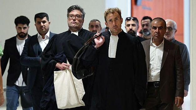 A lawyer is seen walking in a court building with a serious expression in the foreground, wearing his court robes and carrying a leather satchel on his shoulder. In the background, at least seven men in suit jackets - relatives of the victims - are seen also walking with serious expressions.