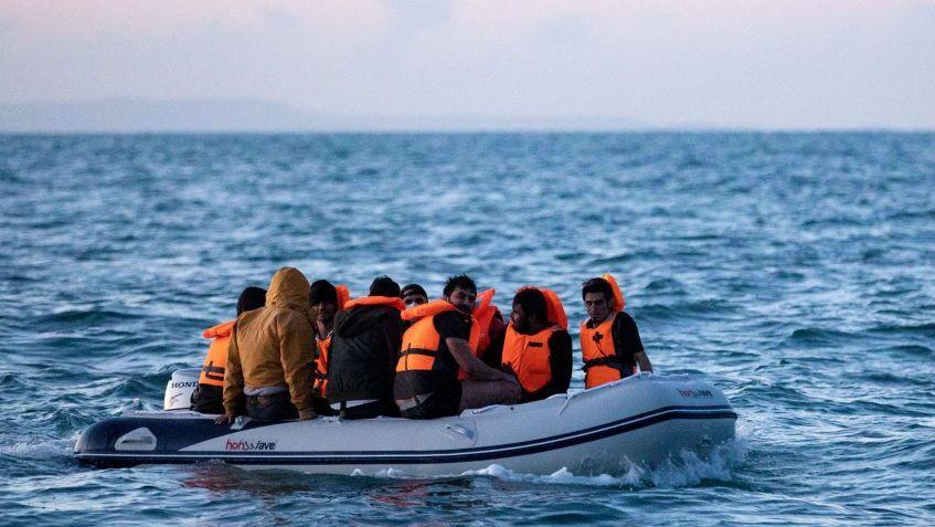 Migrants on a small inflatable boat attempt to cross the English Channel near the Dover Strait, the world's busiest shipping lane. The people on board the dinghy are wearing orange life jackets. The waves are splashing around the vessel. One person without a life jacket is wearing a brown jacket with a hood.