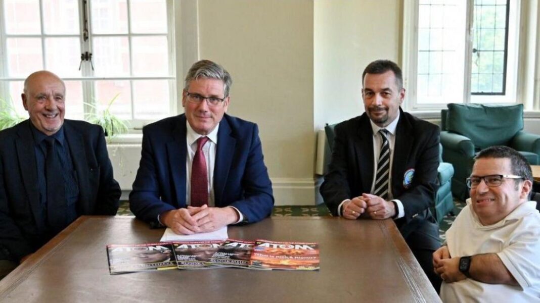Sir Keir Starmer sits in the centre of three campaigners behind a large wooden desk. He is smiling and wearing a navy suit with a white shirt and a red tie, and has his hands clasped over a pile of documents in front of him. To his left is a campaigner with a grey goatee, wearing a black suit over a navy shirt and tie. To his right are two more campaigners, one with a black beard and wearing a black suit over a white shirt with a black and white striped tie. The campaigner furthest left is in a wheelchair and wears a white polo shirt.