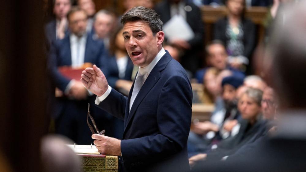 Andy Burnham in the House of Commons at Prime Minister's Questions. He is wearing a dark blue suit, white shirt and blue tie.