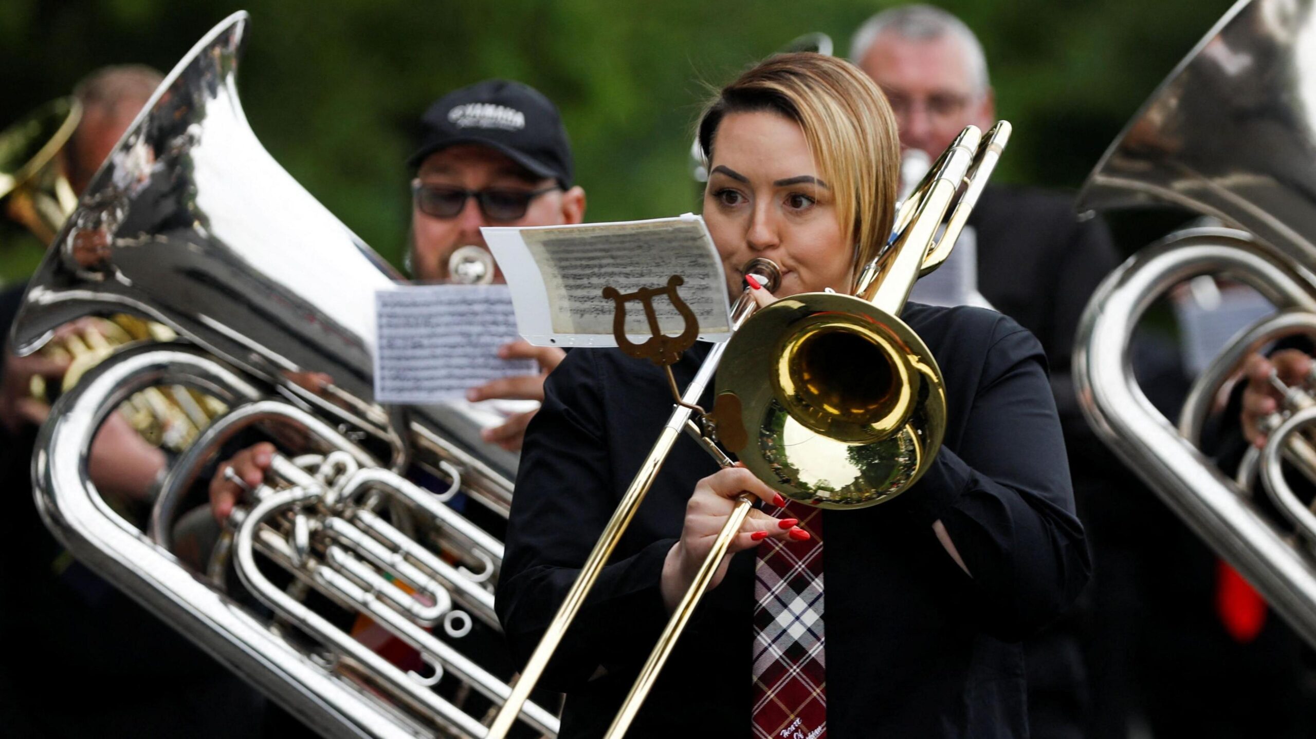 A woman playing a brass instrument. She is a member of a marching brass band which is playing in a parade during the traditional Whit Friday band contest in Manchester in 2024. Behind her can be seen two men who are also playing brass instruments