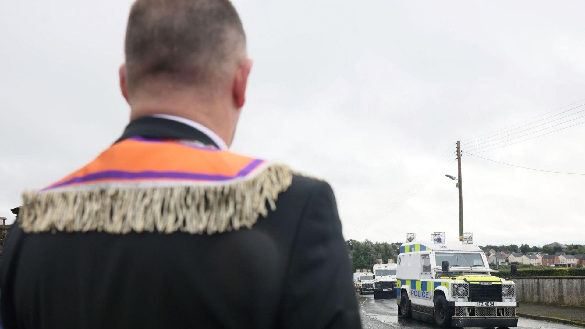 A man is standing looking at a row of police Land Rovers. He has his back to the camera and is wearing an Orange Order sash.