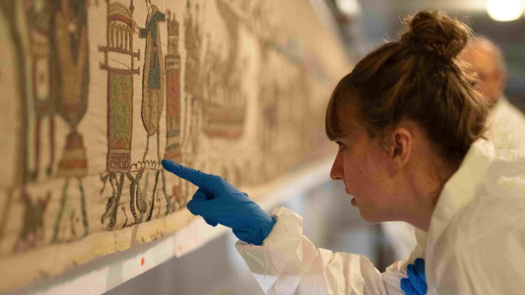A woman in a sterile suit and gloves inspects the artwork with a pointed finger.