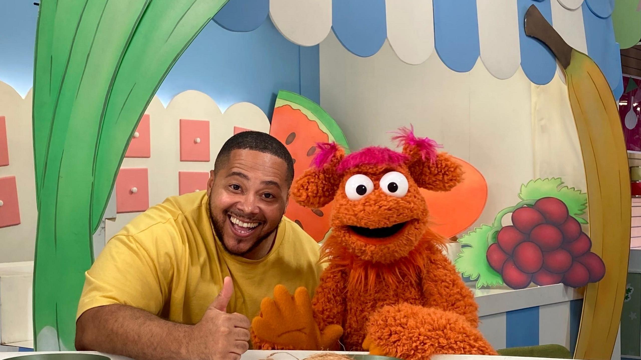 A person in a yellow top sits at a table giving a thumbs-up beside an orange furry puppet with large white eyes and pink hair tufts. The set is decorated with oversized fruit-themed props, including a watermelon slice, raspberries and a banana. Blue-and-white striped panels and colourful market stall graphics create the appearance of a children's television market scene.