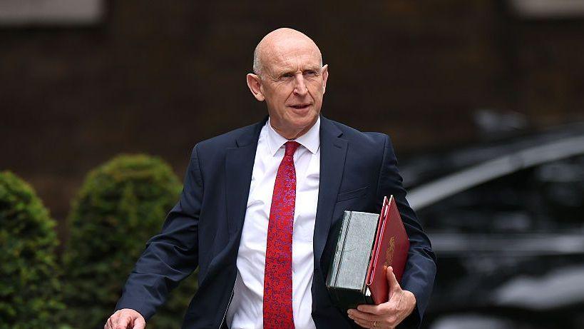 An image of John Healey from the waist up. He is walking and wearing a dark suit with a white shirt and red patterned tie. He is carrying a red folder. The background is blurred by you can see that he is outside from the outline of bushes.