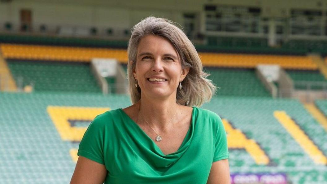 Julia Chapman, who has shoulder-length blonde hair and is wearing teardrop-shaped earrings, and a green top, stands inside Franklin's Gardens stadium at Northampton Saints. She has a wide green.