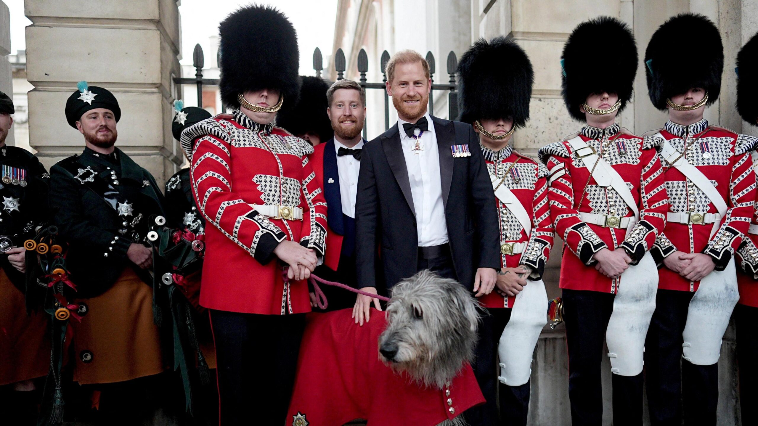 Prince Harry, Duke of Sussex, poses in a black tuxedo with his hand on a large grey dog as members of the Irish Guards' Band and guards dressed in red and white blazers stand behind.