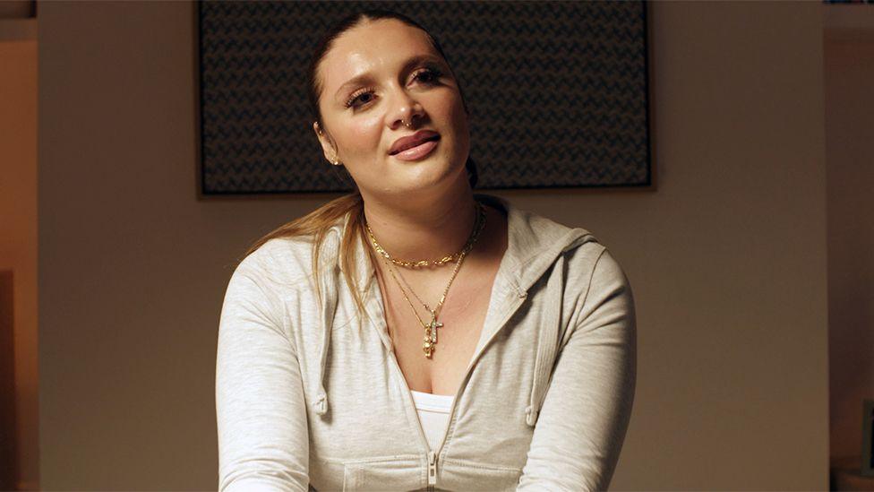 Medium close-up of Violet seated indoors under warm lighting, wearing a light-coloured zip-up hoodie over a white top. Several necklaces, including a cross pendant, are visible. She is facing slightly away from the camera, with a framed patterned artwork on the wall behind her and a softly lit interior background.