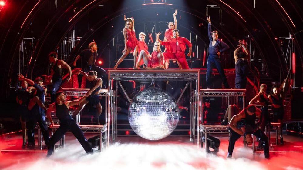 Five red-clad dancers on top of a metal contraption, surrounded by other backing dancers in black.