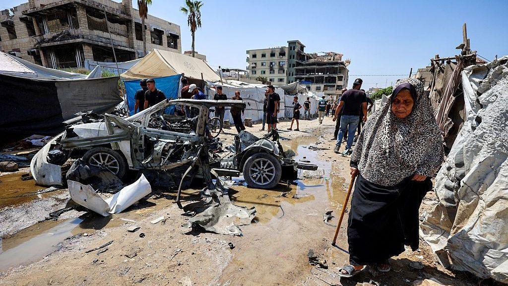 An elderly woman walks past a destroyed vehicle following an Israeli strike in the Rimal neighbourhood of Gaza City (1 September 2026)