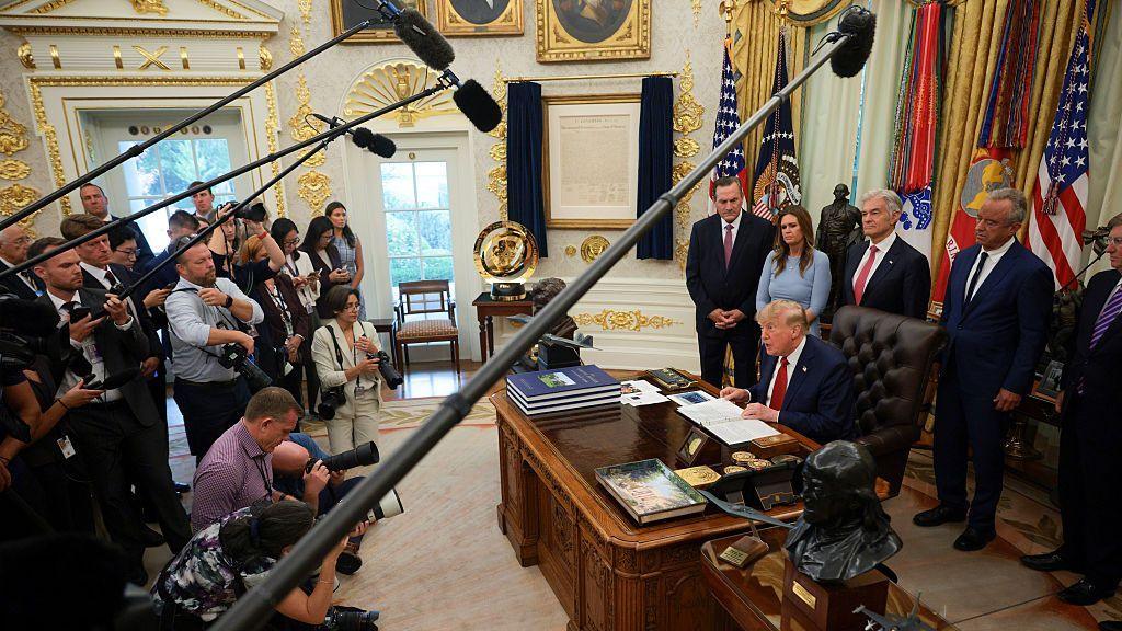 Reporters in the Oval Office with boom mics up to Trump and other officials standing behind him at his desk