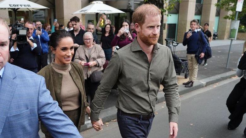 Prince Harry, wearing a khaki shirt and navy trousers, holds Meghan's hand and as they cross a road, protected by a man in a blazer clearing the way for them to walk. In the background are a crowd of people taking photos.