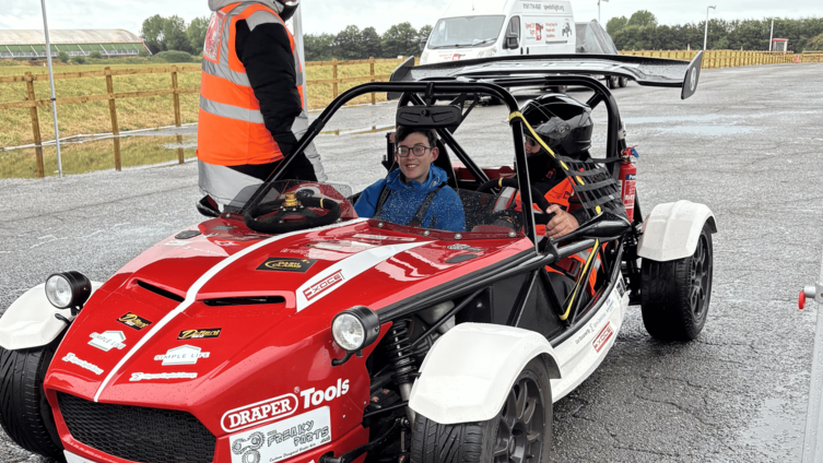A boy in a blue jacket with brown hair and glasses is sitting in a sport car.  Next to him is someone in a helmet.
