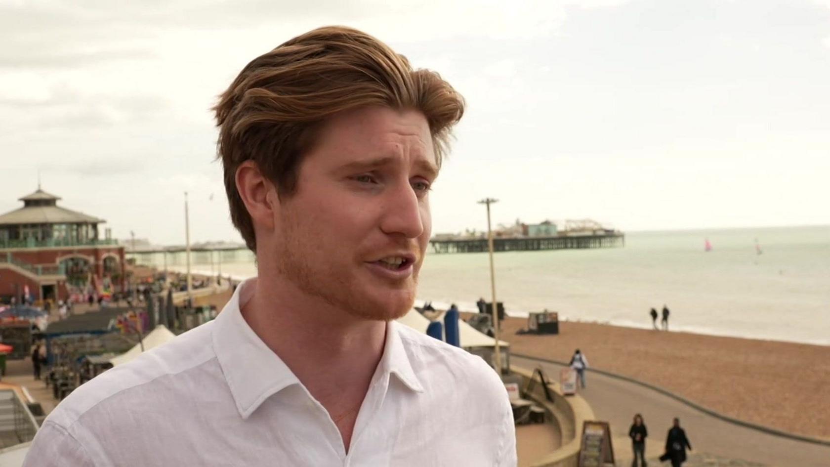 A man in his twenties wearing an open-necked white shirt being interviewed on a balcony overlooking a beach.