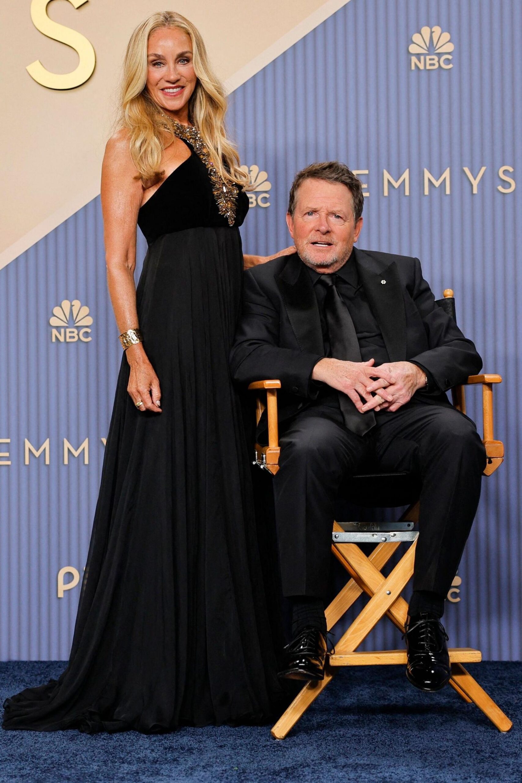 Michael J Fox in a black suit, seated in a director's chair, with Tracy Pollan next to him with one hand on his back