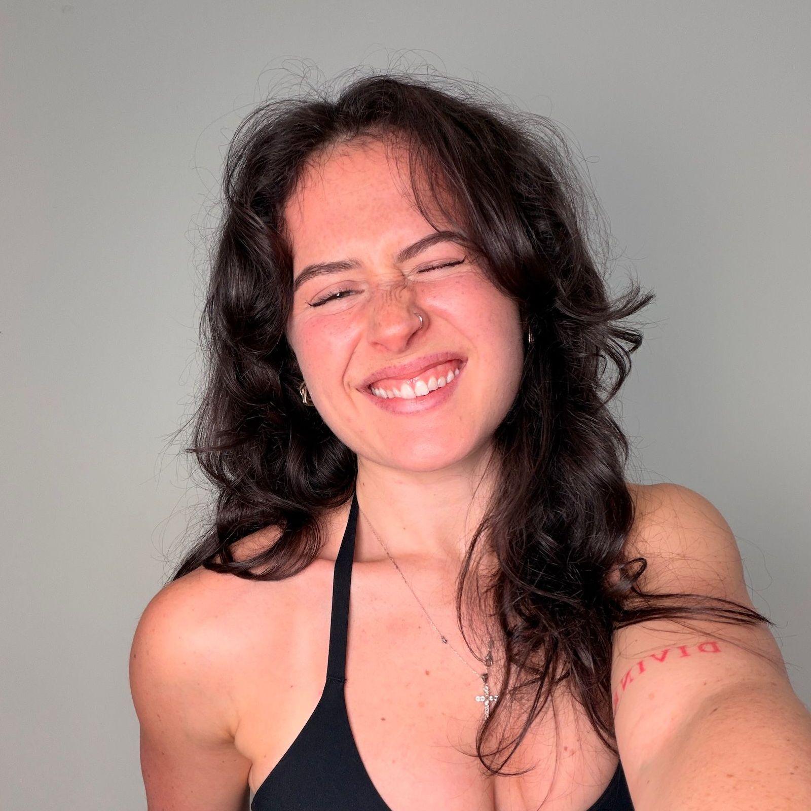 Sarah Mackay smiles and squints at the camera while taking a selfie. She has long dark brown hair and is wearing a black sleeveless top; she's standing against a plain white wall.