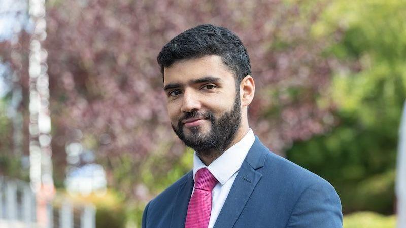 A man with short black hair and a beard is smiling at the camera. He is wearing a blue suit, a white shirt and a pink tie and is standing outdoors with greenery in the background that has been blurred.