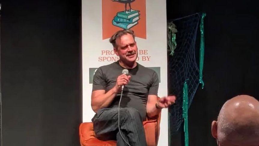 A man sitting with one leg over another in an orange chair. He's holding a microphone and looking at someone in front of him with an inquisitive look. Behind him is a pull-up banner with books on. He appears to be on a stage.