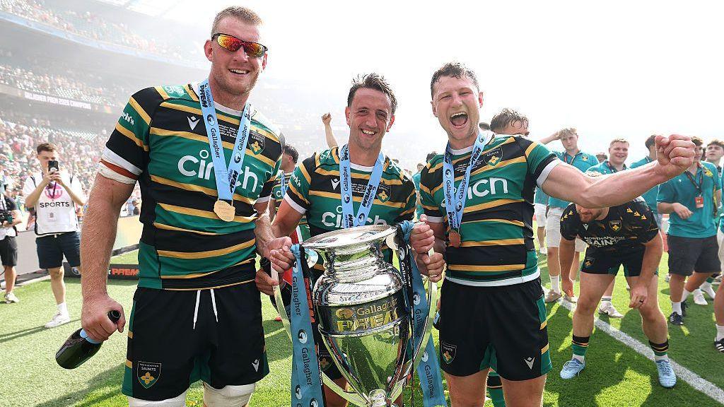 Alex Mitchell standing on Twickenham pitch holding Prem trophy, hot and sweaty wearing home Green, Black and gold kit and flanked by two team mates with othes celebrating into camera behind them