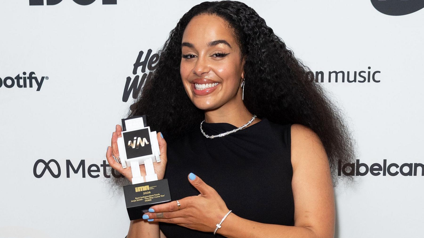 Jorja Smith wears a sleeveless black dress paired with a silver necklace and bracelet. Her nails are painted blue and she holds her award while smiling at the camera. She has long thick black hair which is parted in the middle and sits past her shoulders
