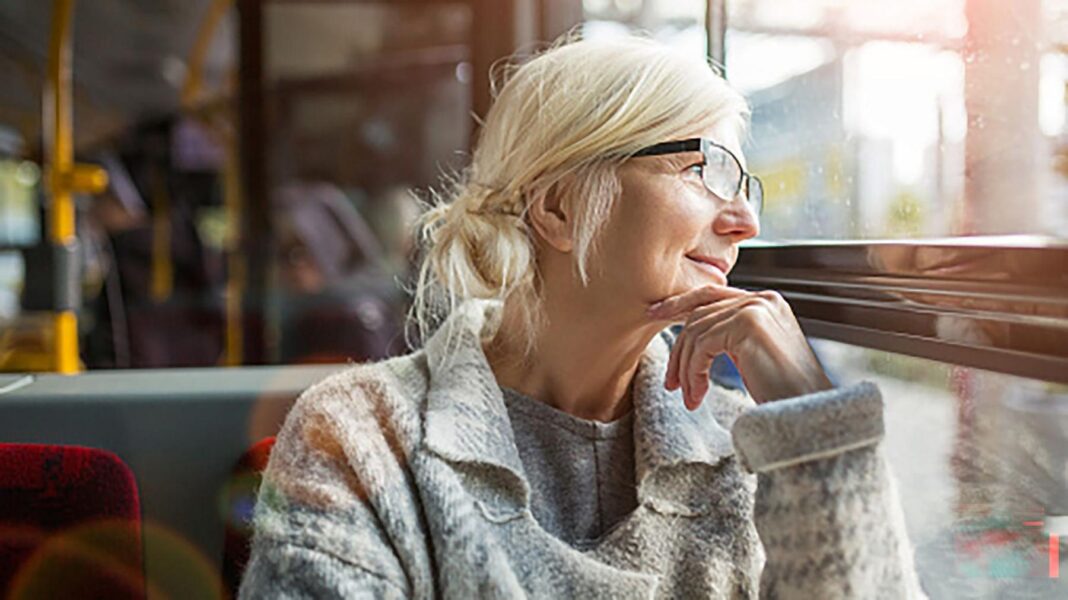 An older woman smiles as she looks out of a bus window. She has short grey in a bun and is wearing a grey jumper.