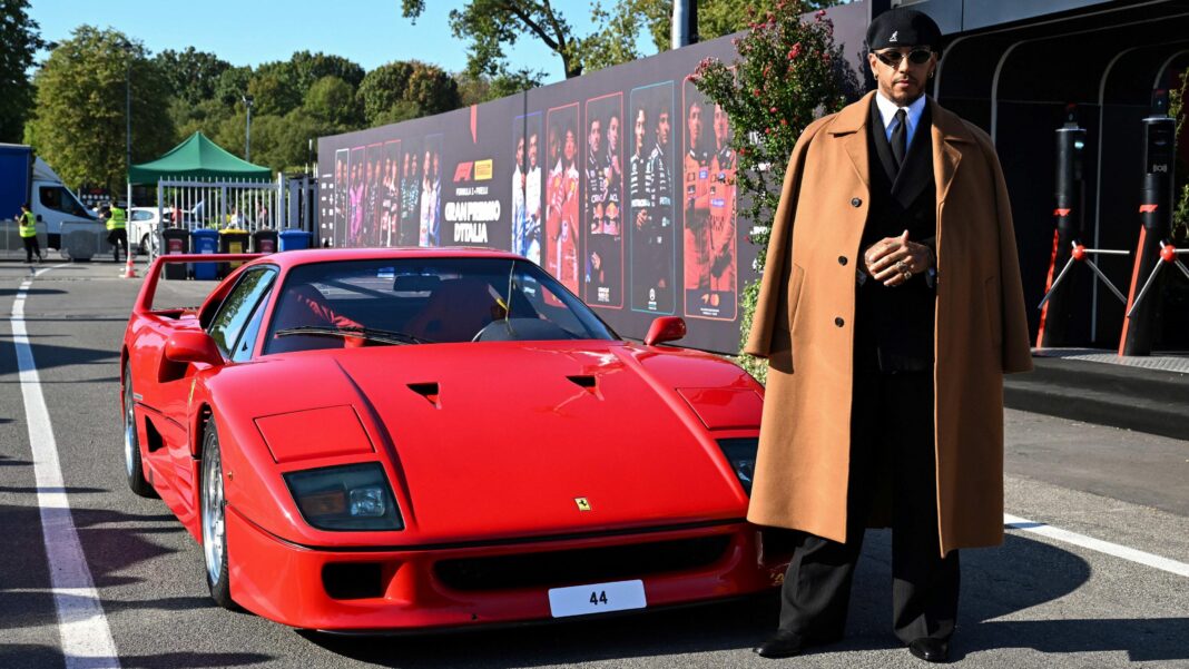 Lewis Hamilton stood next to a red Ferrari F40. He is wearing a brown jacket over a suit and tie