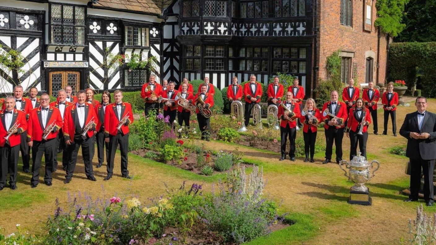 Members of the brass band, dressed in red jackets with the Foden's band logo on their lapels, are spread around the lawned areas of an ornamental garden in front of a black and white half-timber frame building.