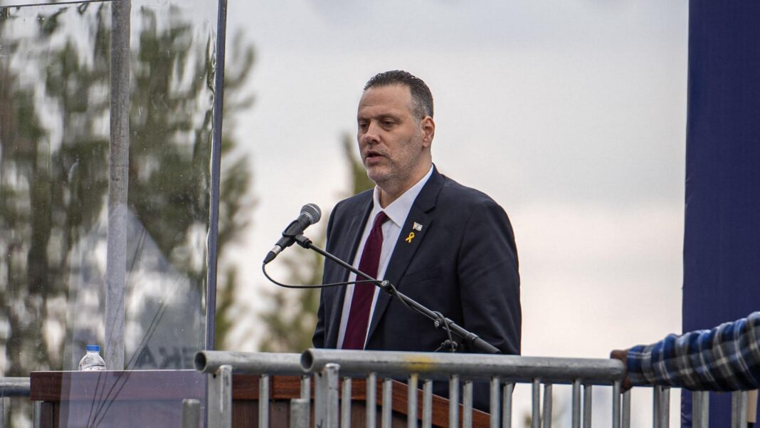 Miki Zohar, wearing a suit and dark red tie, speaks into a microphone on a podium.