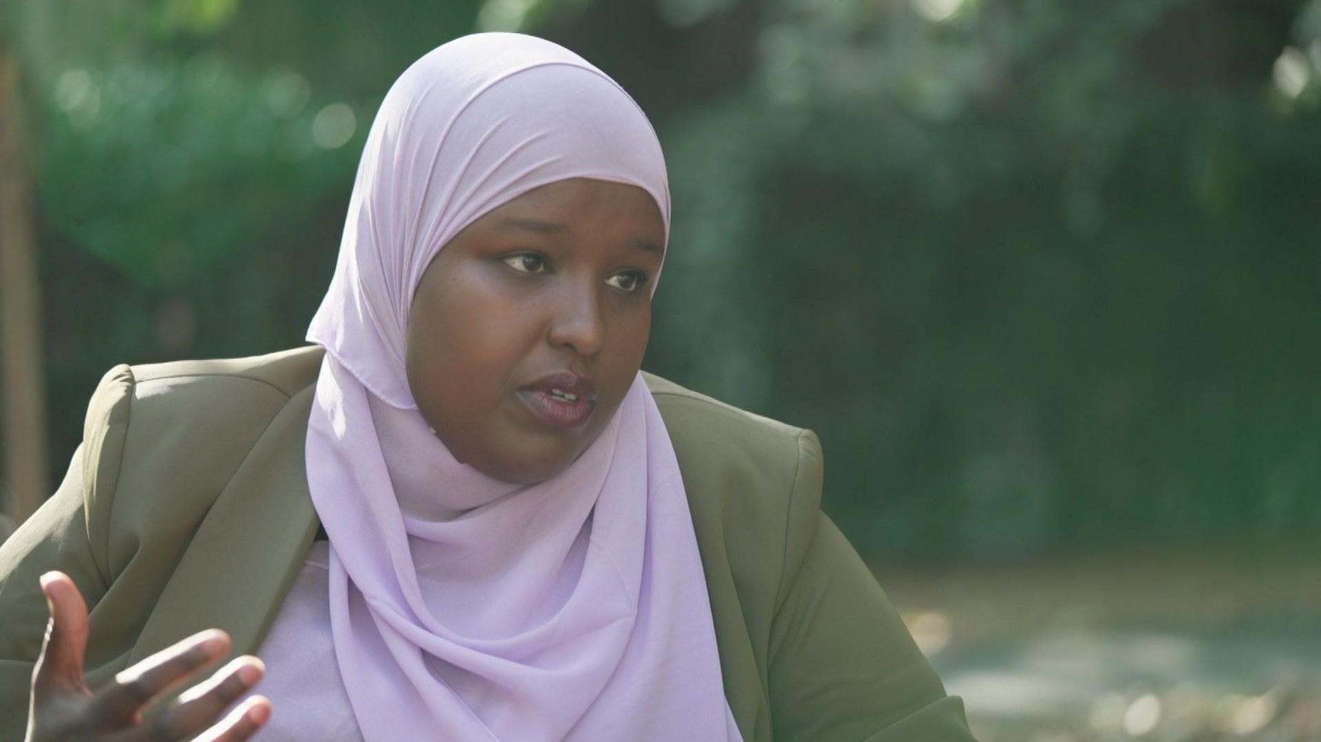 A lady in a pink headscarf and brown/green jacket is sitting for an interview in a park.