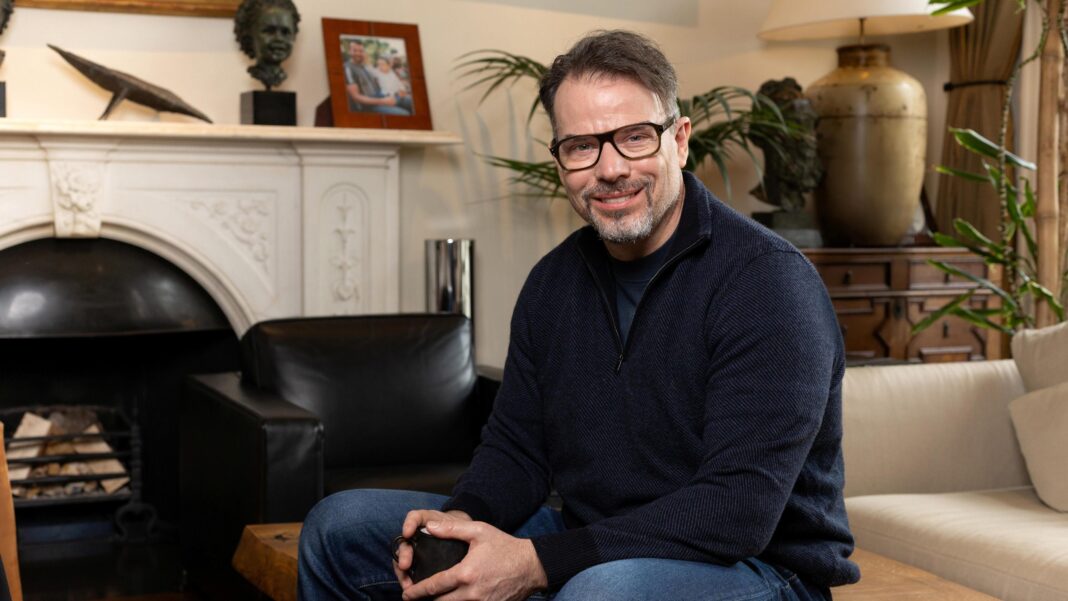 UK’s third-biggest taxpayer to leave for Greece Chris Rokos sitting in a room wearing a dark blue jumper and blue jeans. In the background there is a fireplace, sofa and chest of drawers.