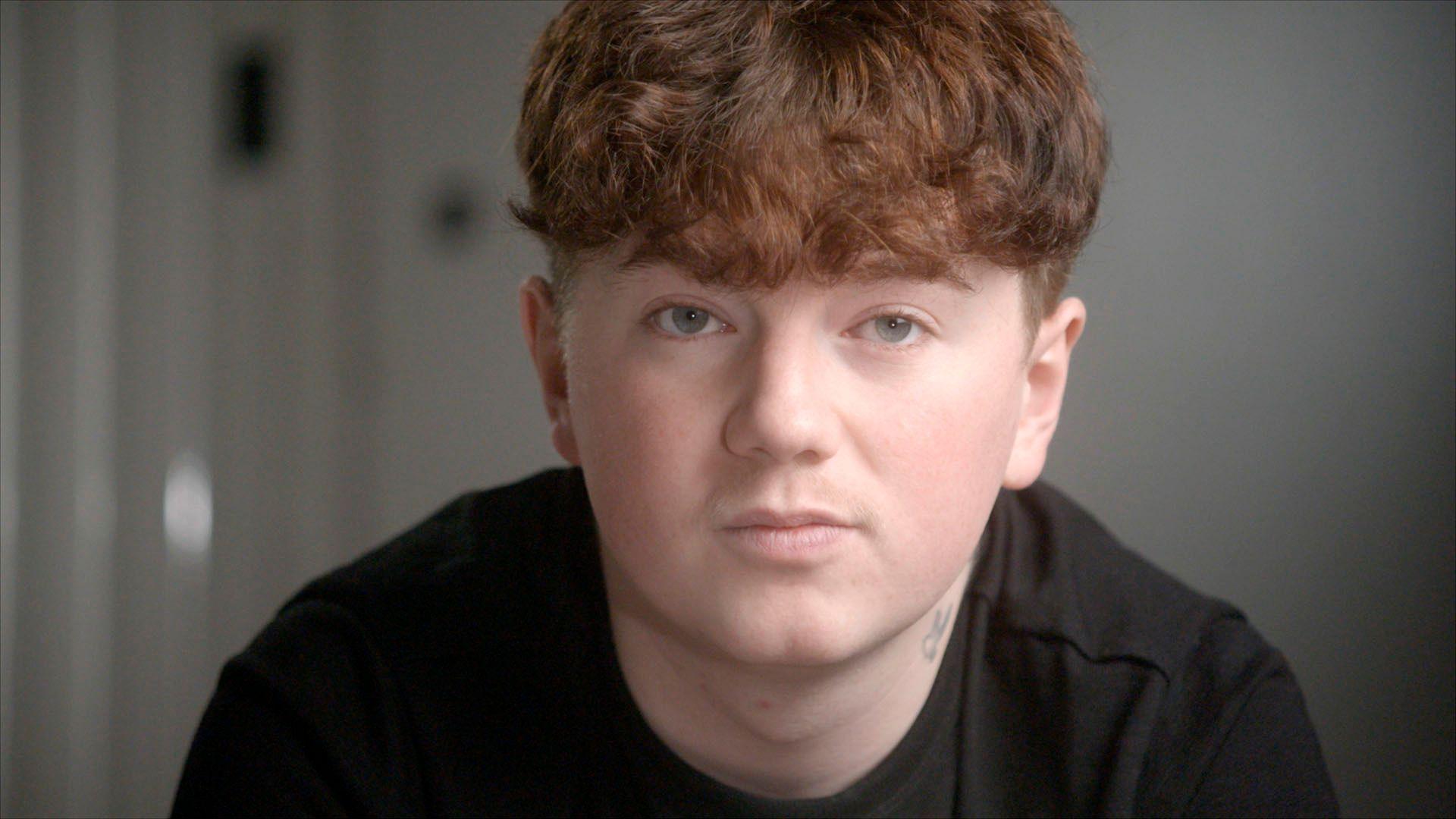 Close-up of Alex's face in the foreground, with short curly ginger hair and a dark top, looking towards the camera. A small neck tattoo is visible above the collar. The background is an indoor setting with a plain wall and a blurred door handle or fixture on the left. No bags, bins, or visible markers are present.