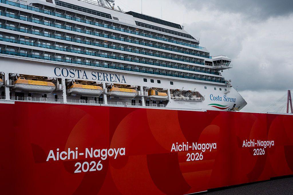 The Costa Serena cruiseliner against cloudy skies, with big boards reading Aichi-Nagoya 2026 in the foreground
