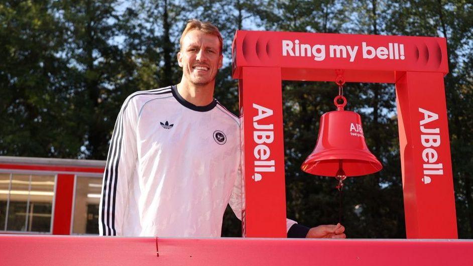 Dan Burn at the start of the Great North Run. He is wearing a white Adidas shirt with the black lines on the sleeve. He stands next to a red start bell with the words Ring my Bell and AJ Bell in white letters.