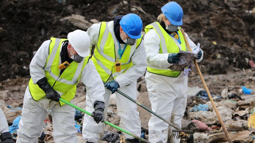 Two men and a woman wearing white overalls, hard hats and yellow high-vis vests searching through a landfill site.