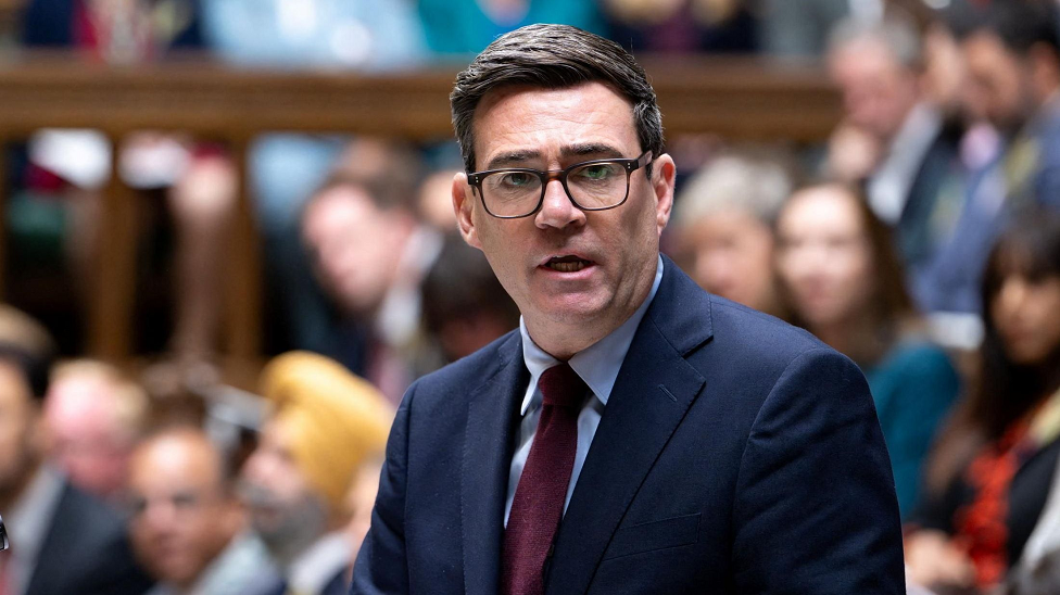 UK Prime Minister Andy Burnham speaks at the House of Commons in London, while wearing a navy suit with a burgundy tie and brown glasses.