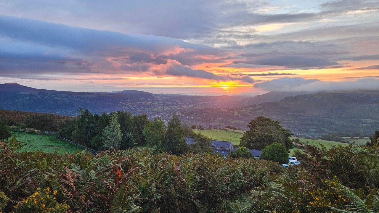 In the distance the sun rises over mountains as a yellow dot in among blue grey clouds. In the foreground are rolling hills and leaves just beginning to change colour