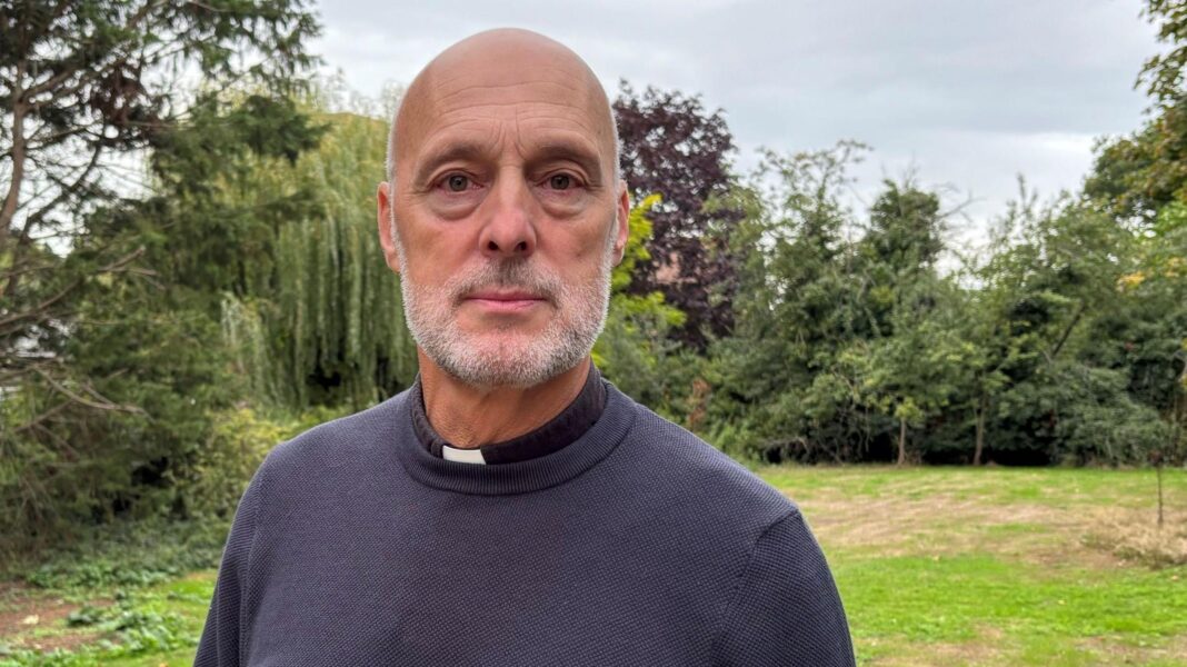 Reverend Chris Willis is standing in a large garden. Behind him is a patch of green lawn bordered with large trees. He is bald with a white beard and is wearing a black sweater.
