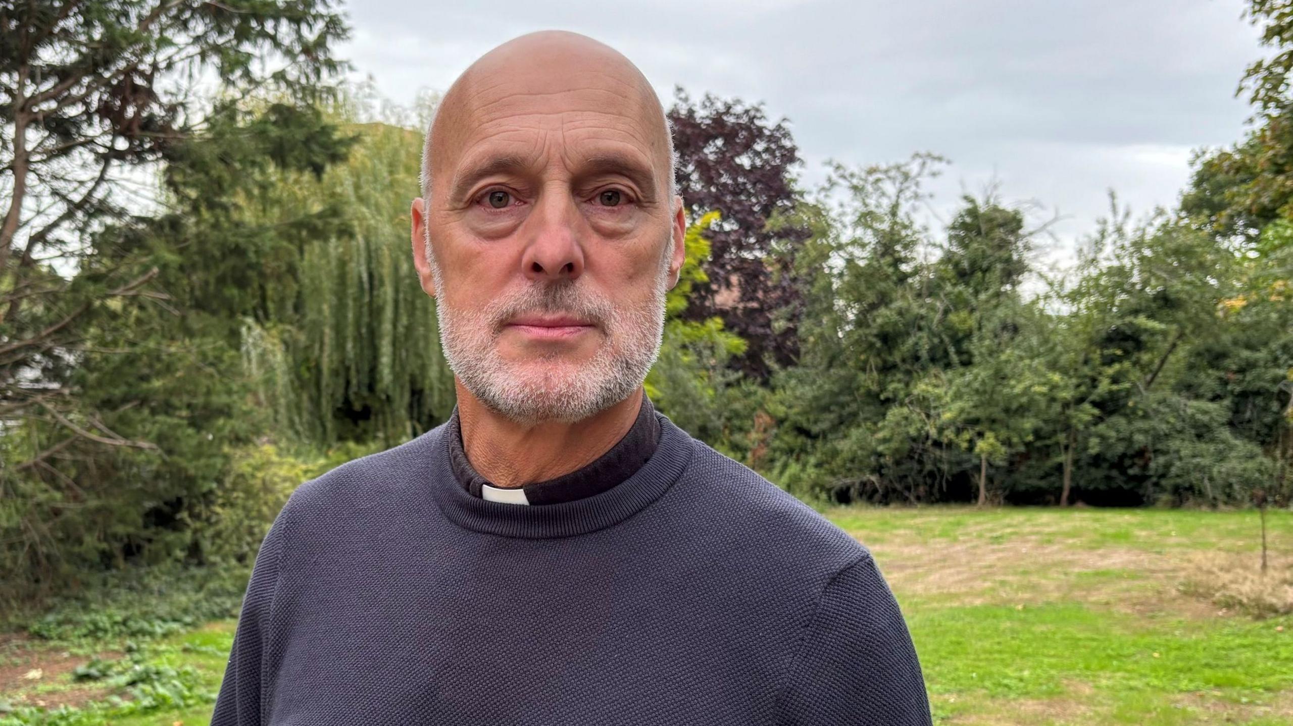 Reverend Chris Willis is standing in a large garden. Behind him is a patch of green lawn bordered with large trees. He is bald with a white beard and is wearing a black sweater.